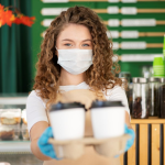 Empleada de cafetería con cubrebocas y guantes sostiene dos vasos de café para llevar en un mostrador, en un negocio de bebidas.