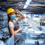 Mujer trabajadora con casco de seguridad y cubrebocas se encuentra en una nave industrial rodeada de estructuras metálicas y equipo de producción.