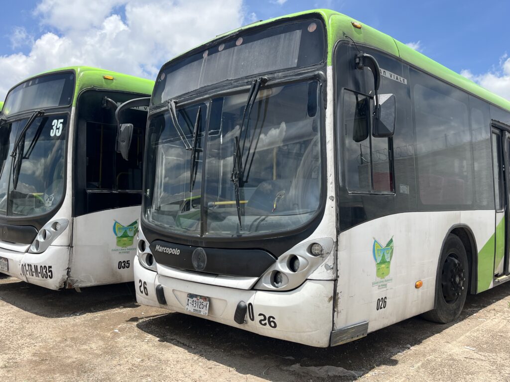 Dos unidades del Tuzobús estacionadas en un patio de resguardo. Los autobuses presentan la cromática verde y blanca del sistema de transporte, con visibles signos de uso, desgaste en la carrocería y algunos daños en la parte frontal, bajo un cielo parcialmente nublado.