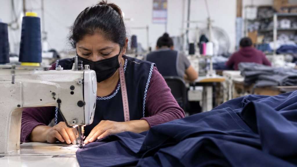 Costurera confecciona una prenda en una máquina de coser industrial dentro de un taller de confección, mientras otras trabajadoras elaboran ropa al fondo; imagen ilustrativa de la industria del vestido en Hidalgo.