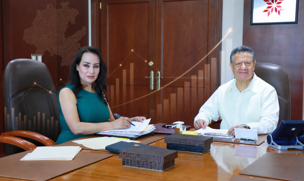 Julio Menchaca Salazar y Esther Ramírez Vargas durante una reunión de trabajo, en una composición ilustrada con gráficas de crecimiento y el mapa de Hidalgo.